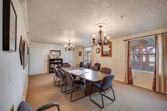 A rectangular activity/dining room with a long table surrounded by chairs, chandeliers overhead, bookshelves, and windows with curtains.