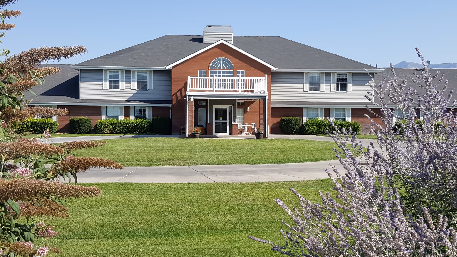 Front exterior view of a two-story senior living facility building with a combination of brick and siding. The building has multiple windows, a small balcony above the entrance, and a covered porch with two chairs. The foreground features a well-maintained lawn and flowering bushes on both sides.