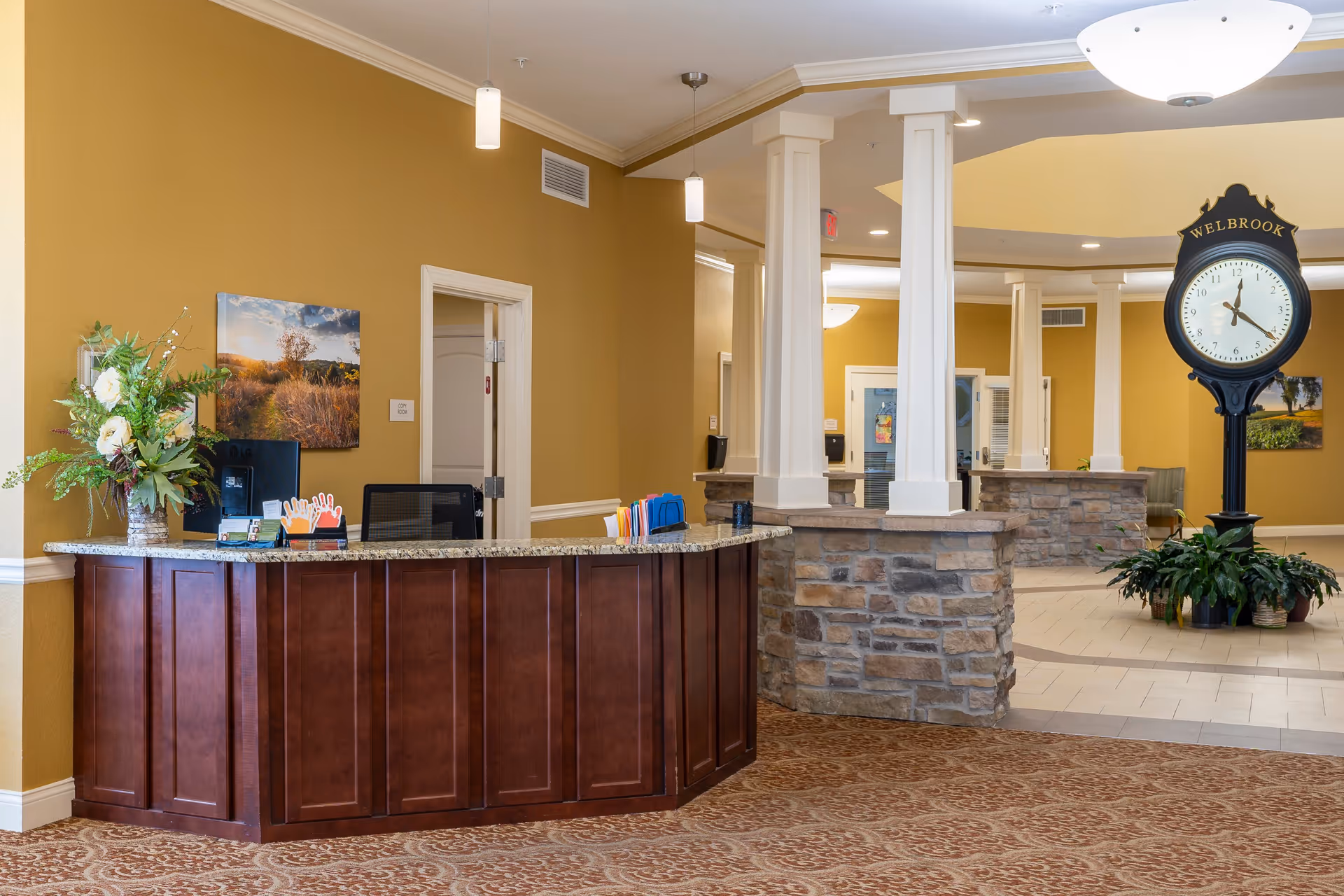 Reception area with a wooden front desk topped with granite, a computer, and a flower arrangement. Behind the desk is a doorway and a wall with a landscape painting. The area features stone pillars and a large black clock labeled 'WELBROOK' surrounded by plants. The walls are painted mustard yellow and the floor is carpeted with a patterned design.
