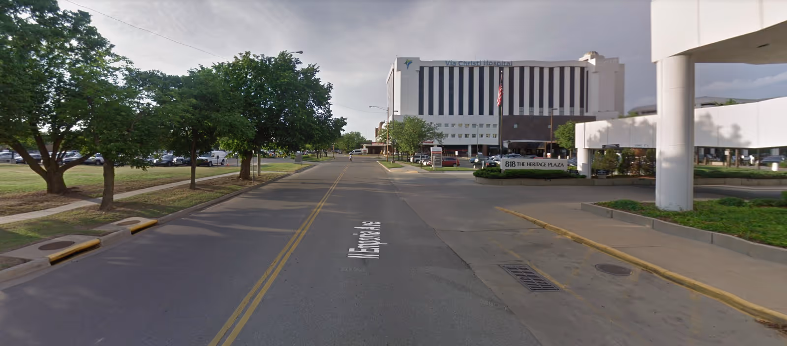 Street view of a hospital-like building across the road, with trees and a sidewalk.