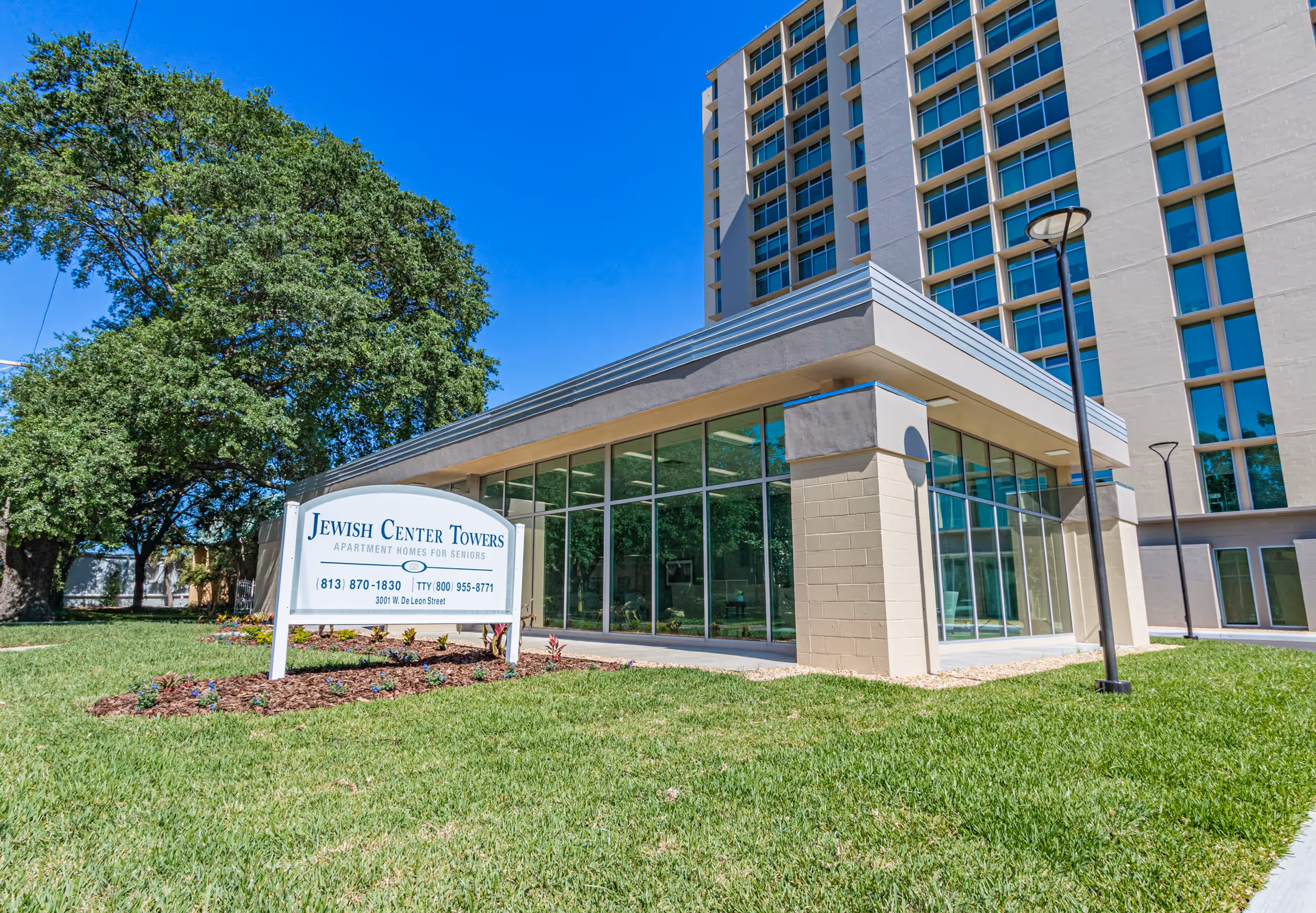 Front exterior of the Jewish Center Towers senior apartment building with a lawn, entrance pavilion, and a sign.