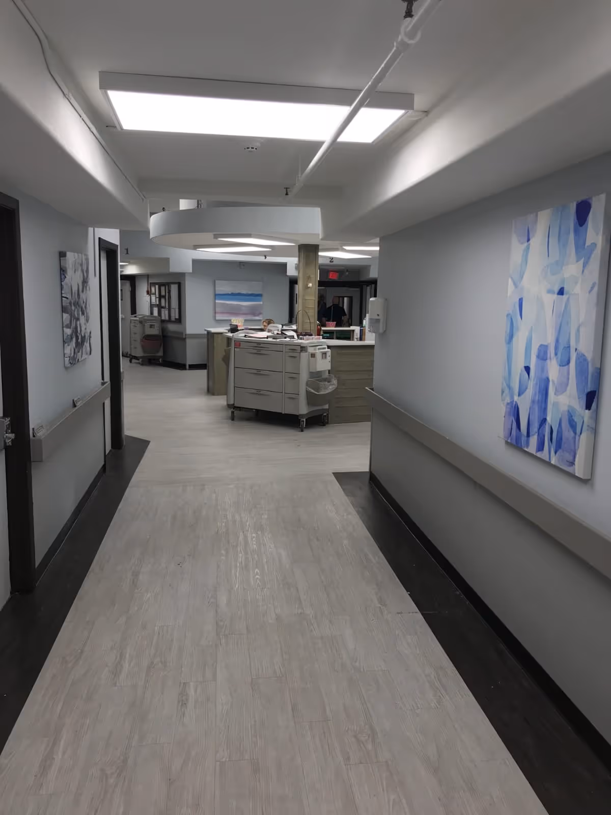 A clean, well-lit hallway in a senior living facility with light wood flooring and gray walls. The hallway leads to a nurses' station with medical carts and equipment. Abstract blue and white paintings hang on the walls.