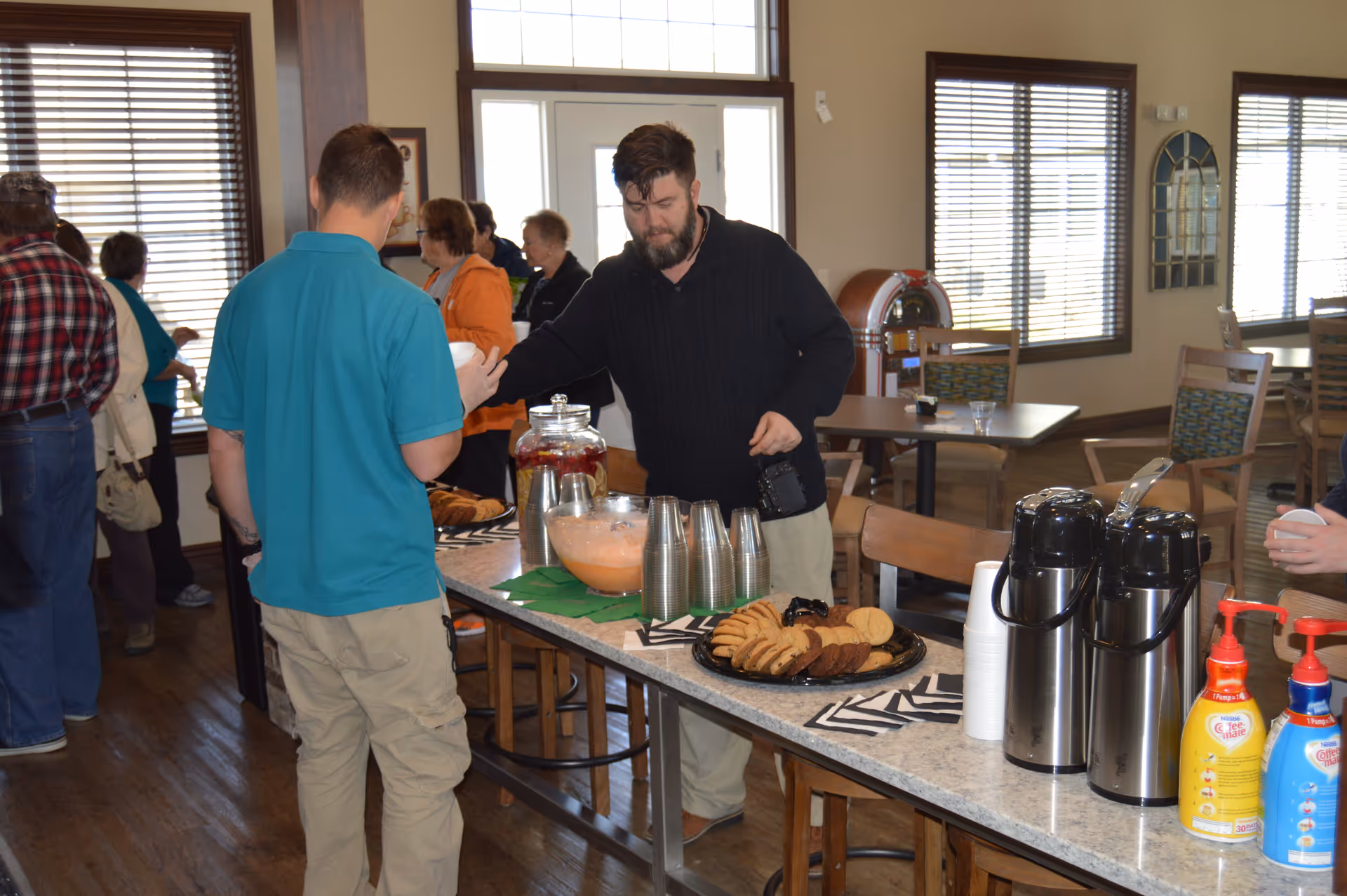 People gather around a buffet table with coffee dispensers, cookies, and punch in a bright communal dining area.