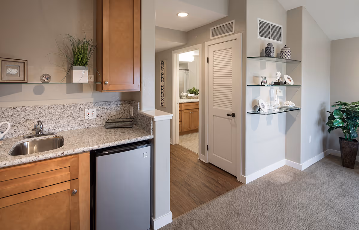 Compact kitchenette with granite counters, wooden cabinets, sink, and doorway to a bathroom.