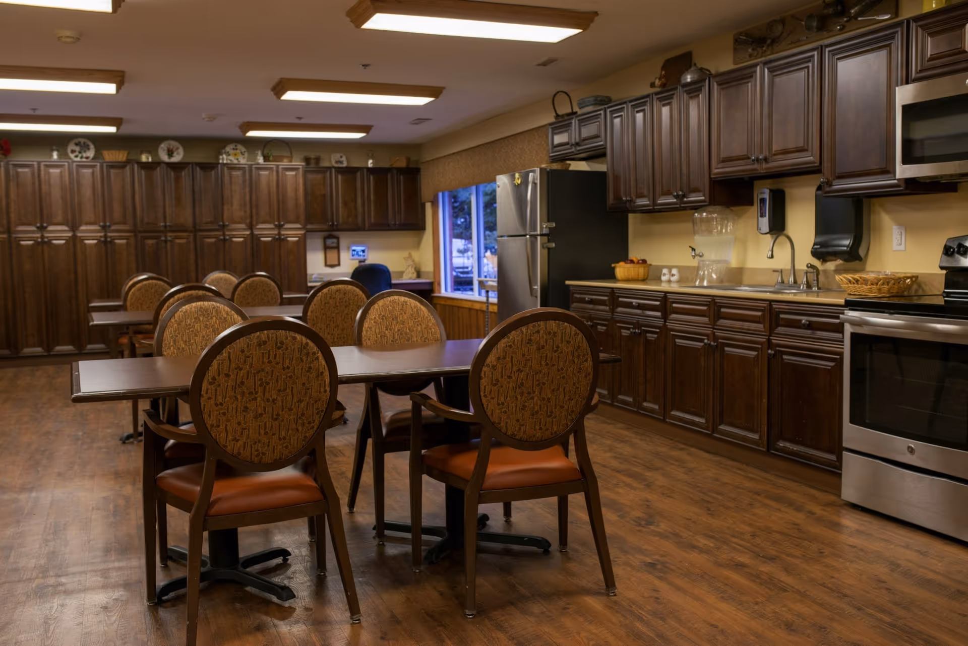 Interior view of a senior living facility kitchen and dining area at Salem Crossing, featuring wooden cabinets, stainless steel appliances, multiple tables with cushioned chairs, and a window with a view outside.
