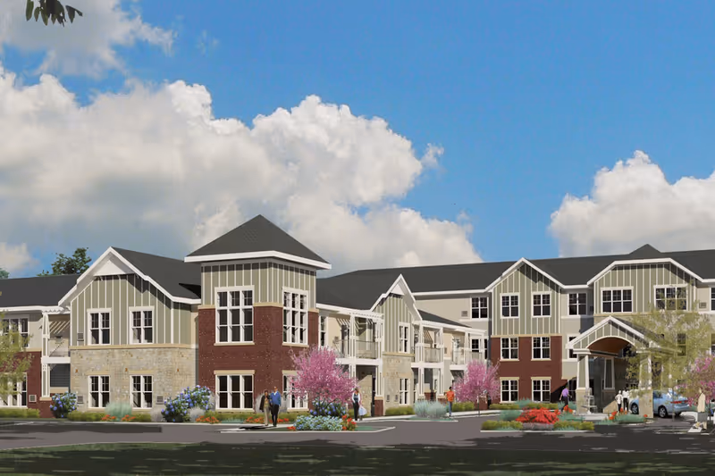 Exterior view of a two-story senior living facility building with a mix of brick, stone, and siding. The building features multiple windows, a covered entrance, landscaped grounds with colorful flowers and trees, and a few people walking near the entrance under a partly cloudy blue sky.