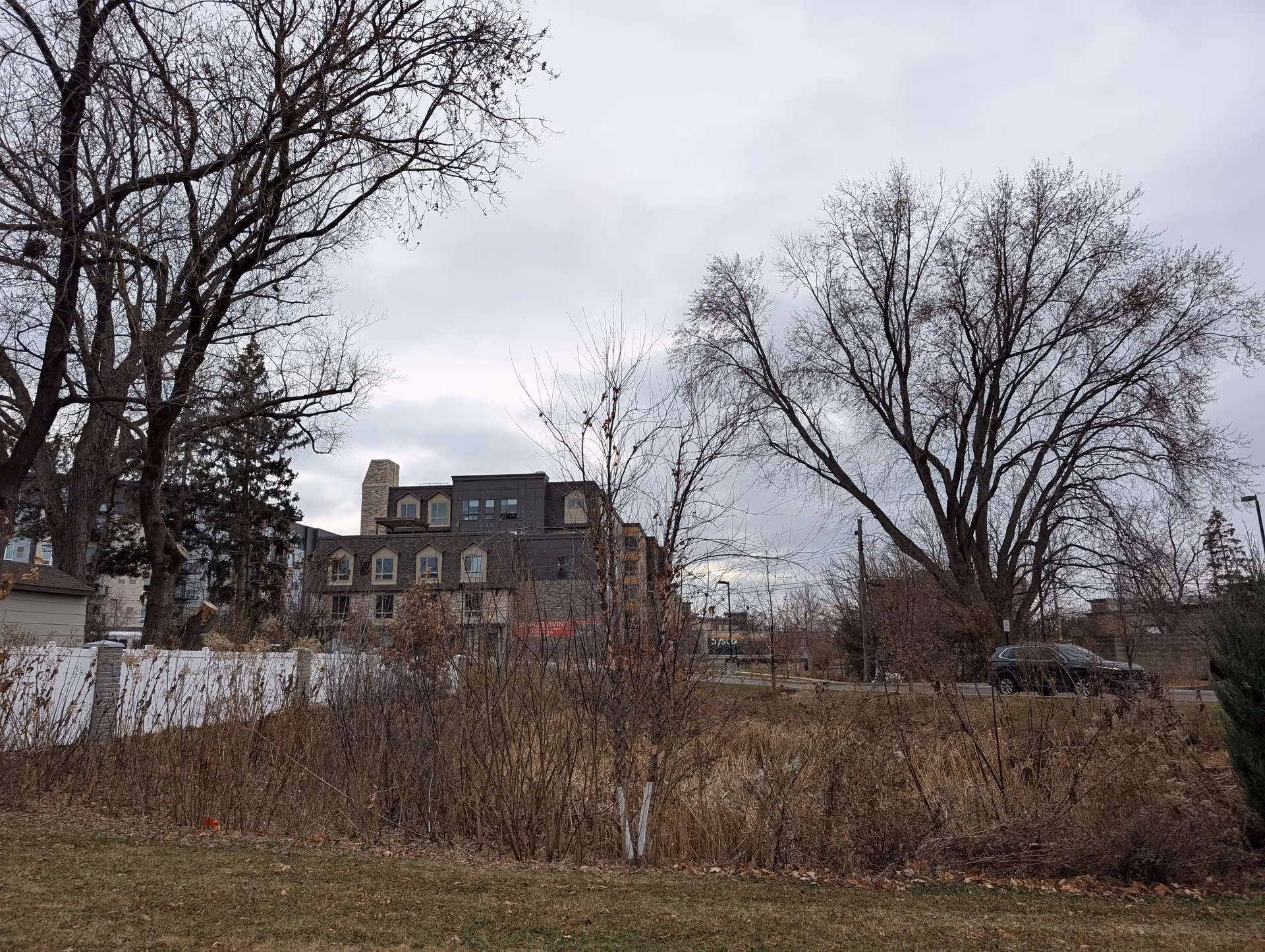 Exterior view of a multi-story senior living building seen through bare trees and brush under an overcast sky.