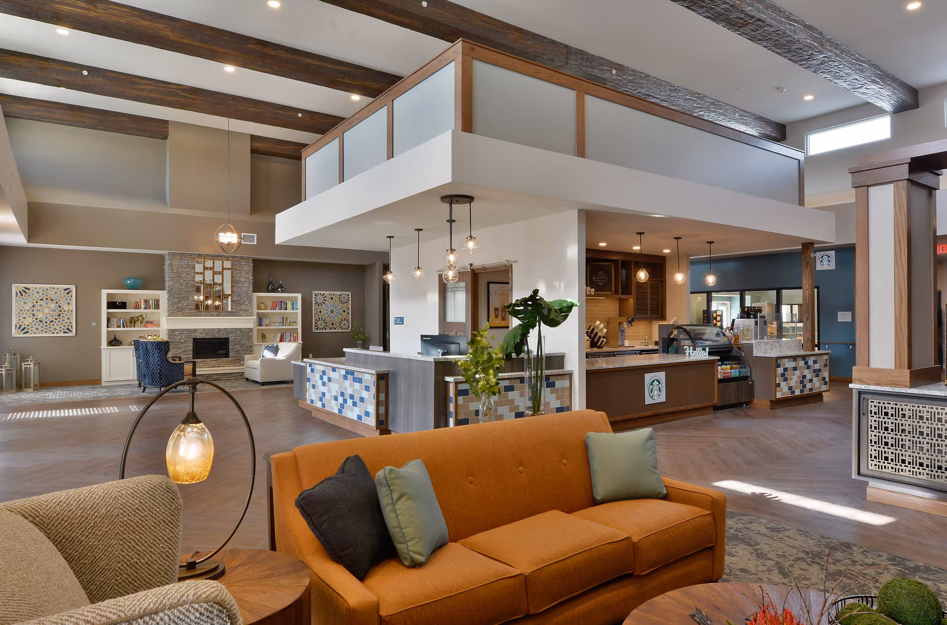 Spacious senior living lounge with sofas and armchairs, a fireplace and bookshelves, and a reception/coffee bar under exposed wooden beams.