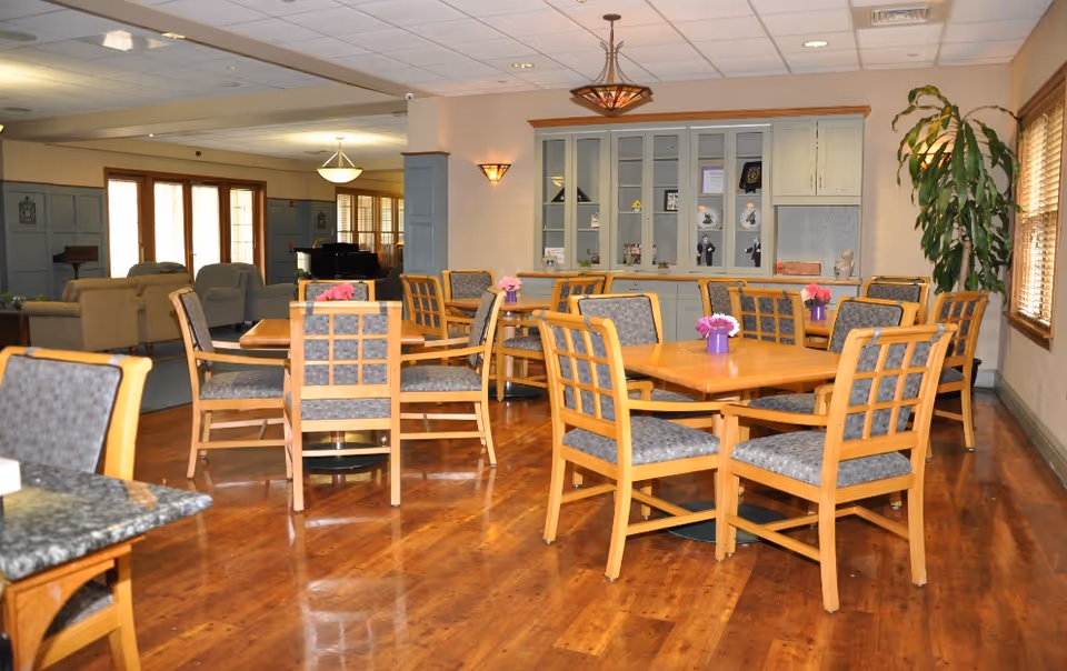 Spacious senior facility dining room with wooden tables and upholstered chairs, small flower centerpieces, and a seating area in the background.