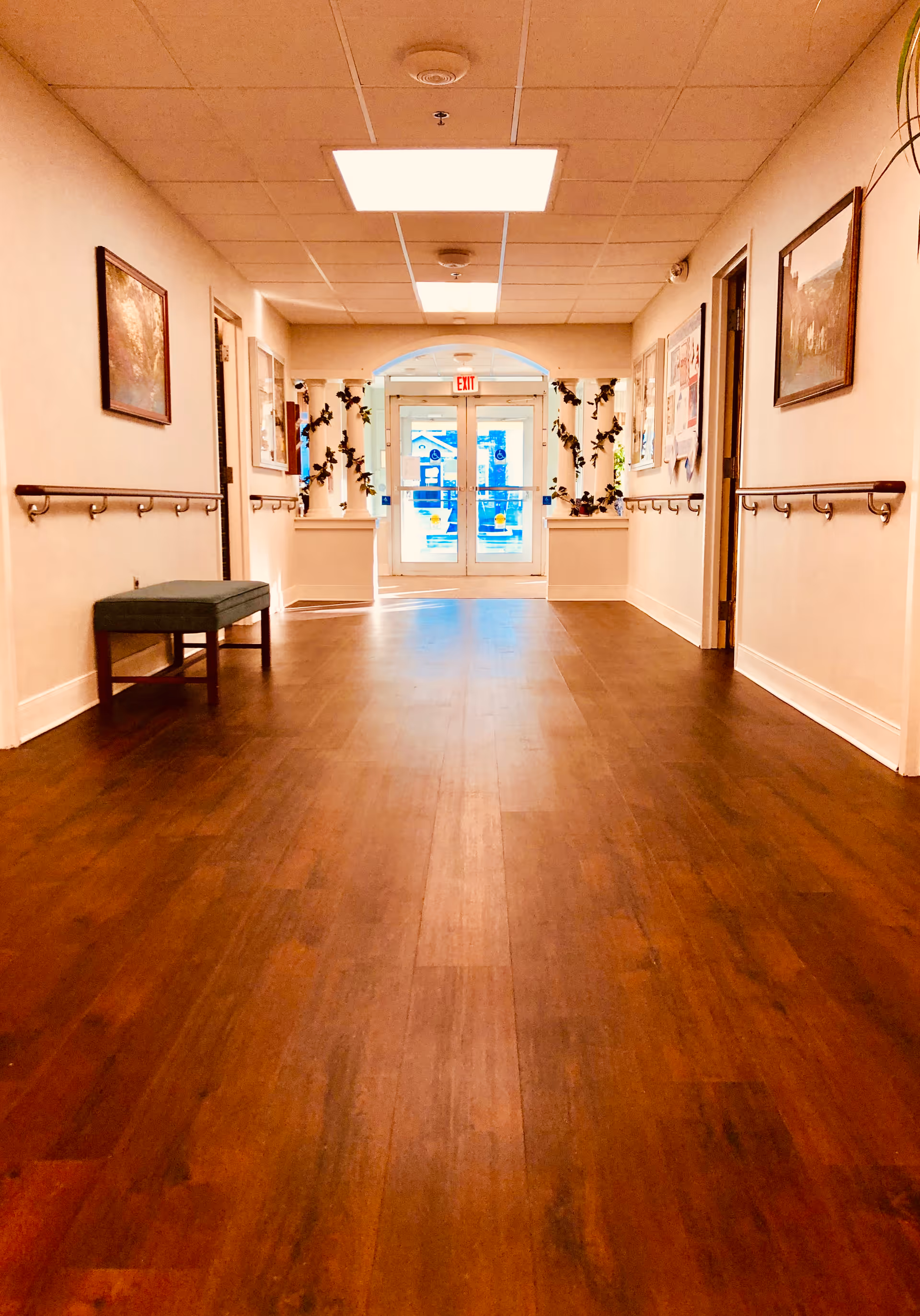 A well-lit hallway with wooden flooring and beige walls, featuring handrails on both sides, framed pictures on the walls, a green bench on the left, and double glass doors at the end with an exit sign above. There are decorative plants around the door area.