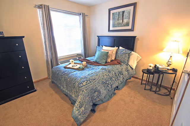 Sunlit bedroom with a double bed in blue paisley bedding, a black headboard, dresser, round side table with a lamp, and a window with curtains.