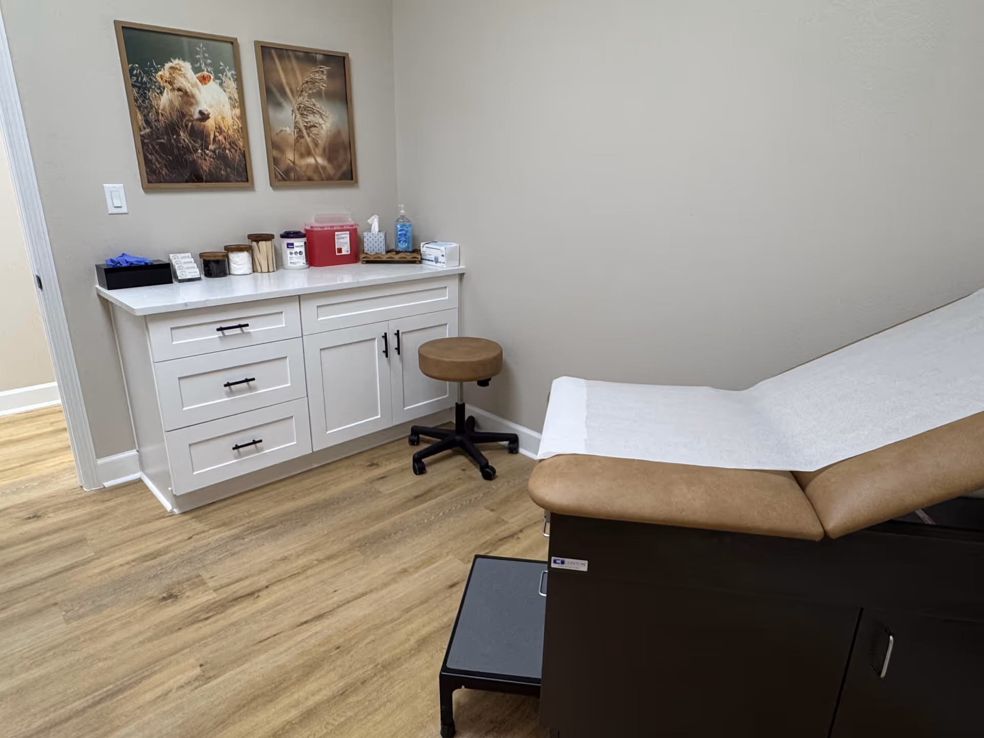 Medical examination room with white cabinet, stool, and examination bed in beige walls.