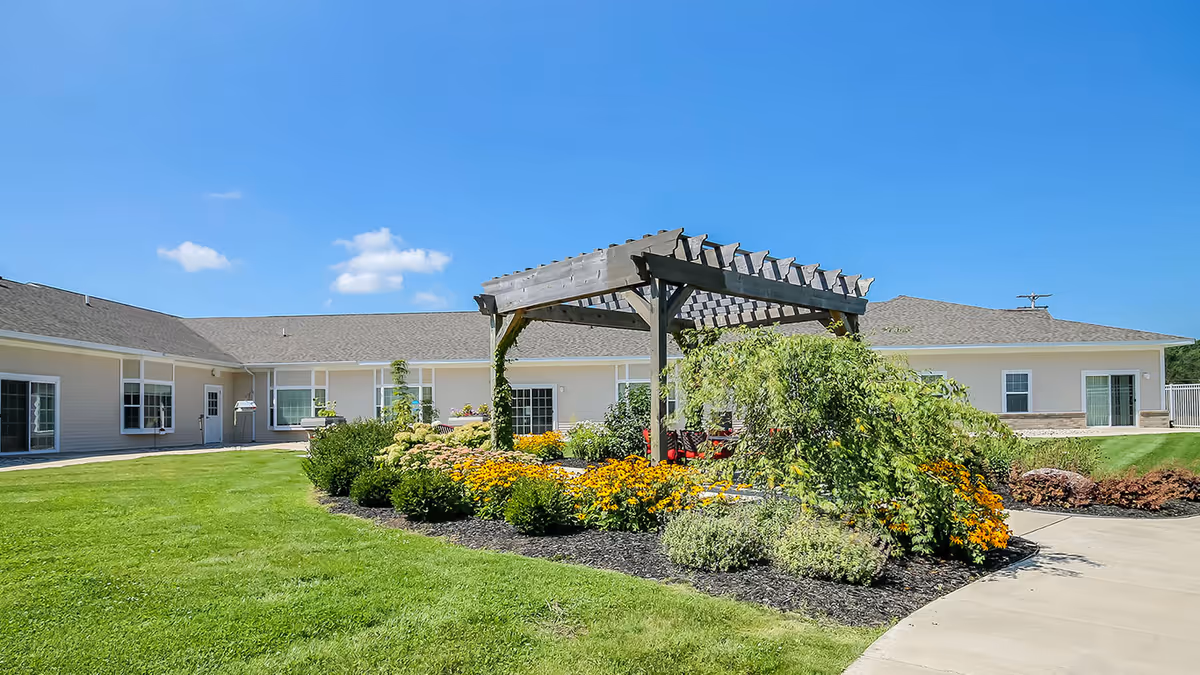 Outdoor garden area at Lakeview Terrace Assisted Living featuring a wooden pergola surrounded by vibrant yellow and green flowering plants, with a well-maintained lawn and a single-story beige building in the background under a clear blue sky.