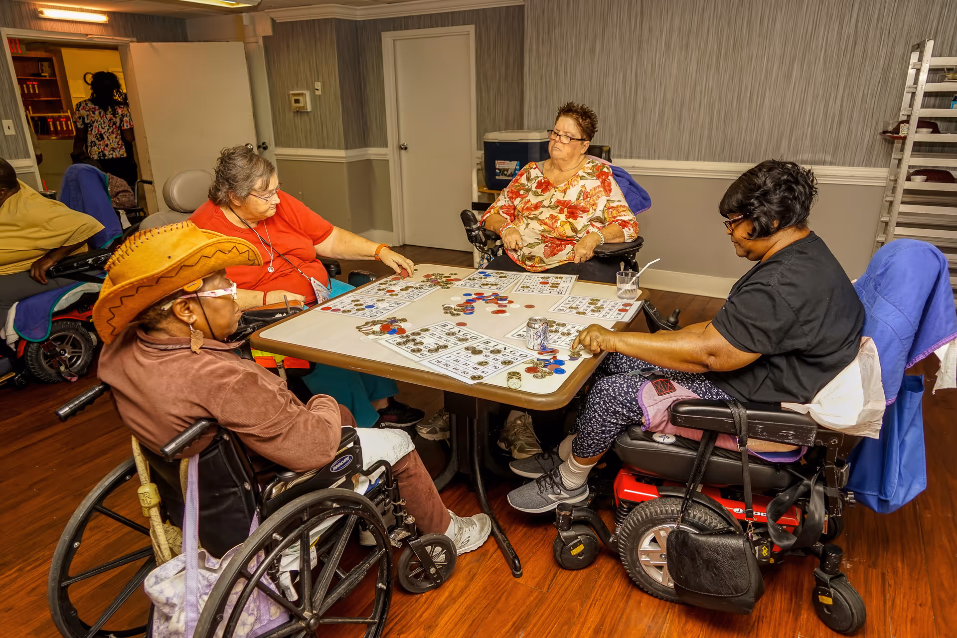 Four elderly women, three in wheelchairs, sitting around a table playing bingo with cards and chips in a room with wood flooring and gray walls.
