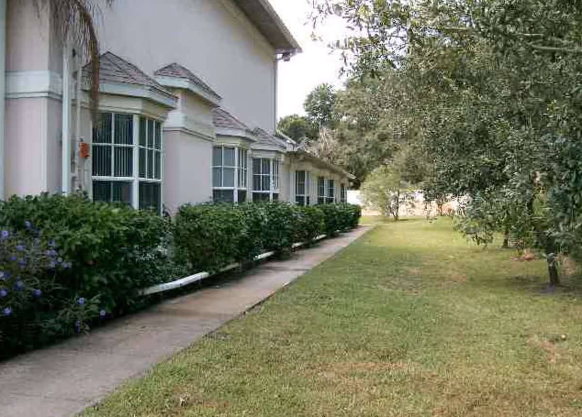 Side view of a building with multiple windows and a sidewalk running alongside it, bordered by green bushes and a grassy area with trees on the right side.