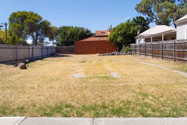 Empty fenced backyard with brown grass, concrete border, and neighboring houses under blue sky.