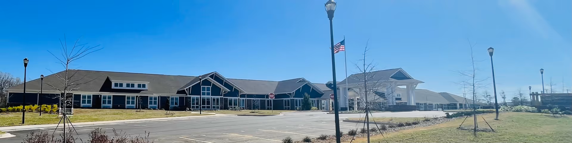 Wide exterior view of a single-story senior living facility building under a clear blue sky, with a large parking lot in front, several young trees planted around, and an American flag on a tall lamp post near the entrance.