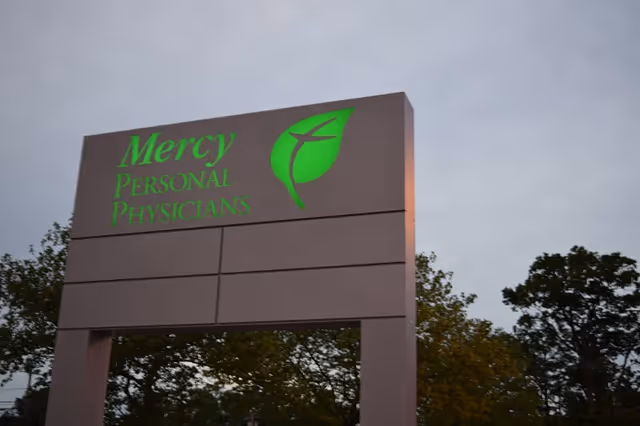 Outdoor signboard reading Mercy Personal Physicians with a green leaf logo against a cloudy sky.