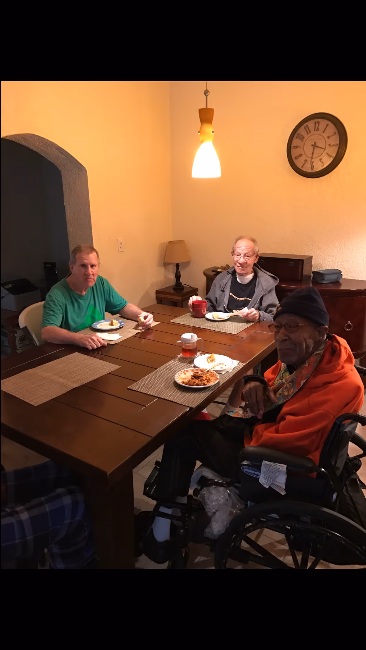 Three older adults sit around a wooden dining table eating a meal in a warmly lit dining room.