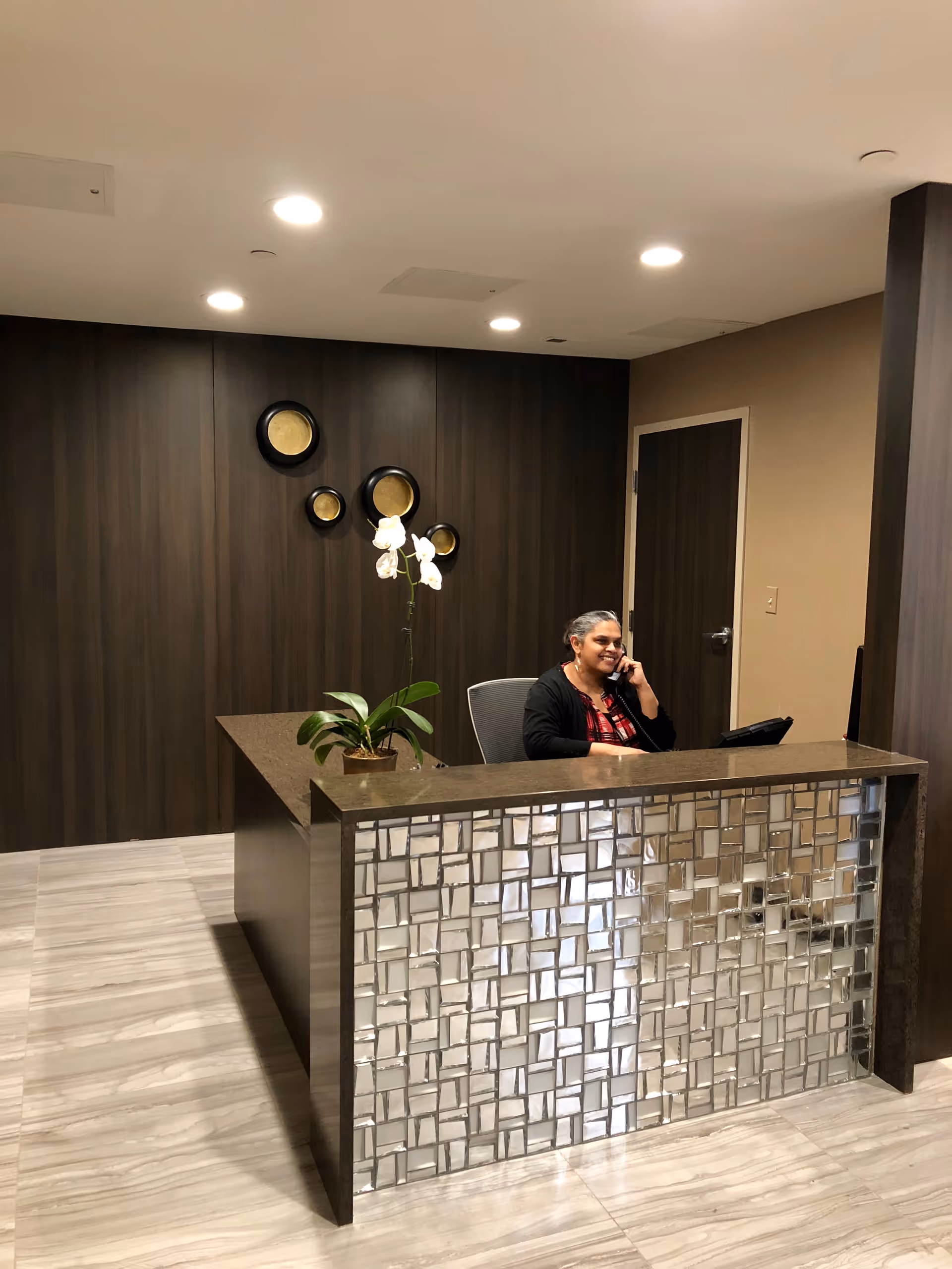 A receptionist sitting behind a modern front desk with a decorative mirrored front panel, talking on the phone. The background features a dark wood-paneled wall with circular wall decorations and a door to the right. A potted orchid plant is placed on the desk.