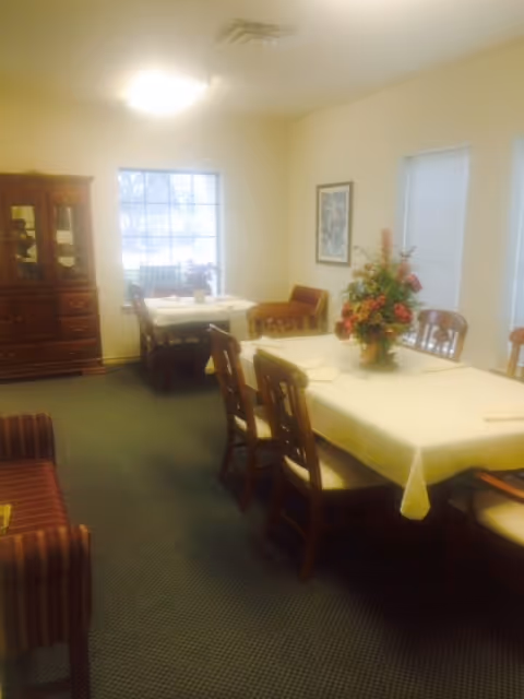 A dining room with two tables covered with white tablecloths, each surrounded by wooden chairs. One table has a floral centerpiece. There is a wooden cabinet with glass doors on the left side and two windows with blinds on the right wall. A framed picture hangs on the wall between the windows.