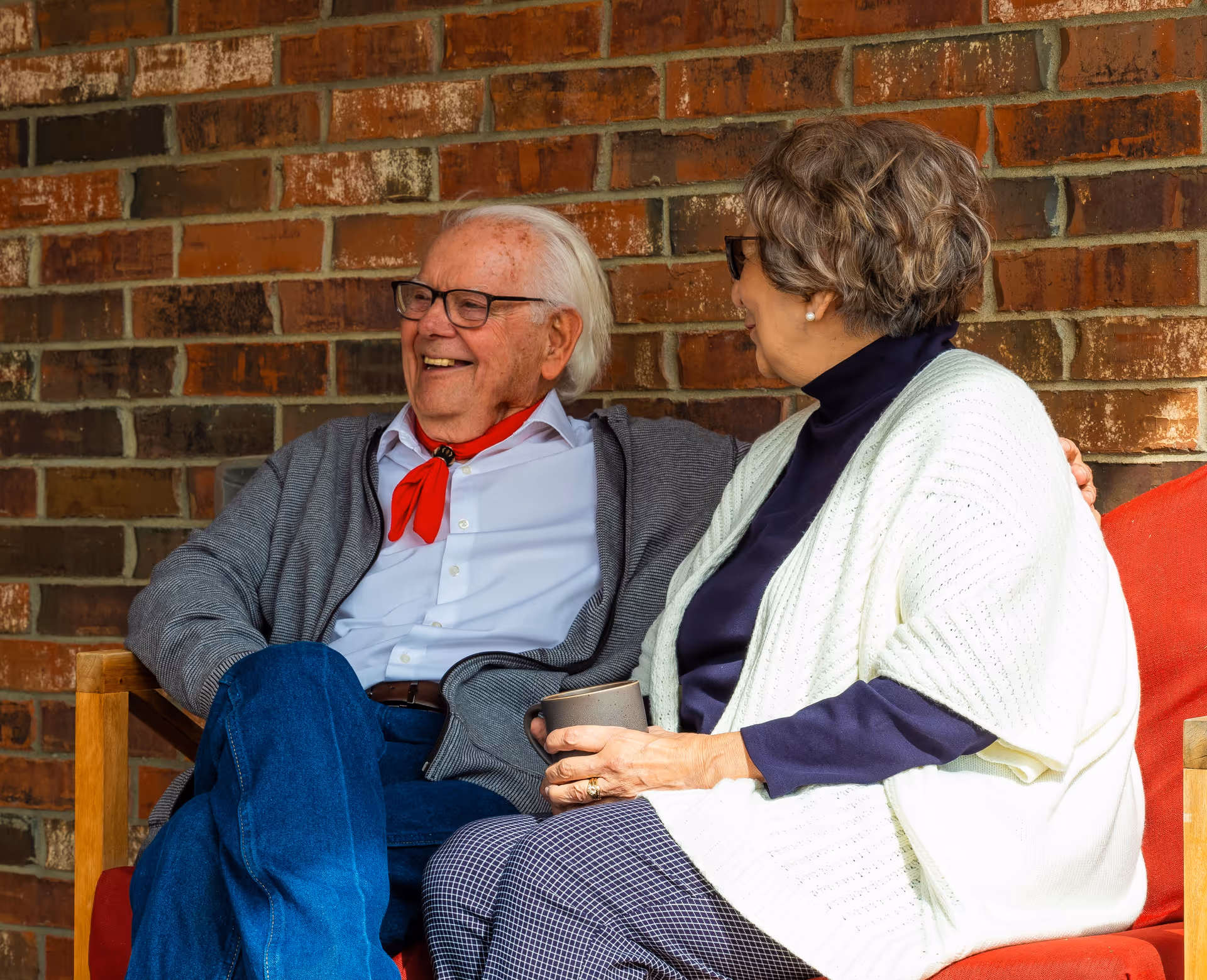 Two elderly people sit on a brick outdoor seating area, smiling and chatting.