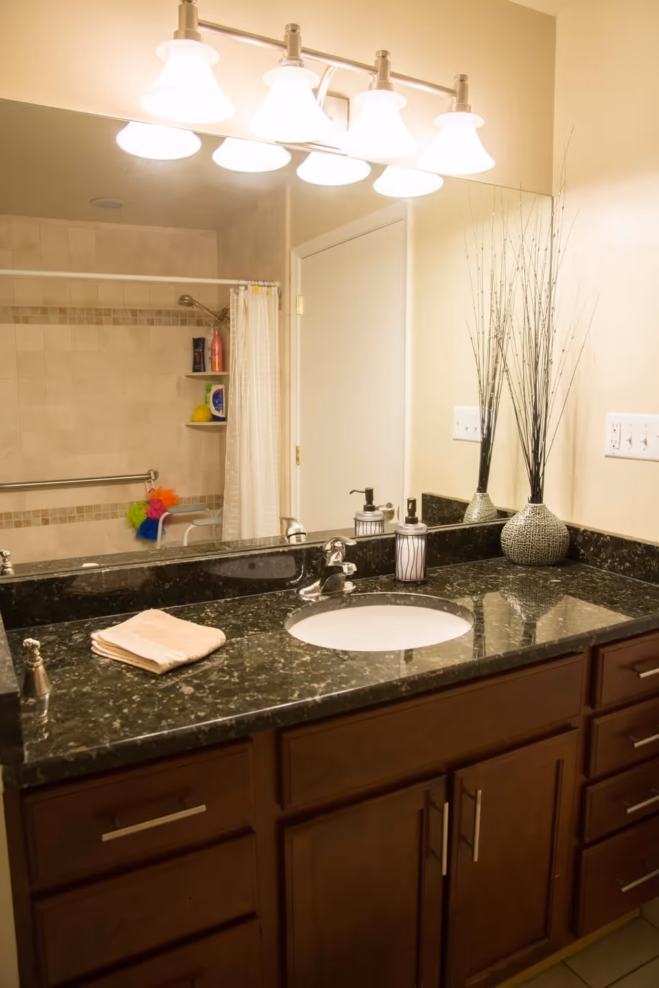 Bathroom vanity with a dark granite countertop, sink, mirror and overhead lights, with a shower visible in the mirror reflection.