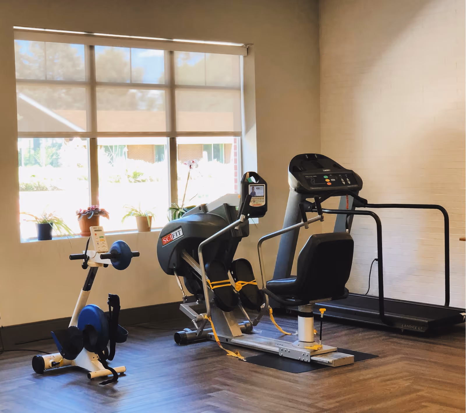 A small exercise room with three pieces of fitness equipment including a recumbent bike, a leg press machine, and a treadmill. There is a large window with a roller shade partially down, and several small potted plants on the windowsill. The floor has a wood-like finish and the walls are light-colored.