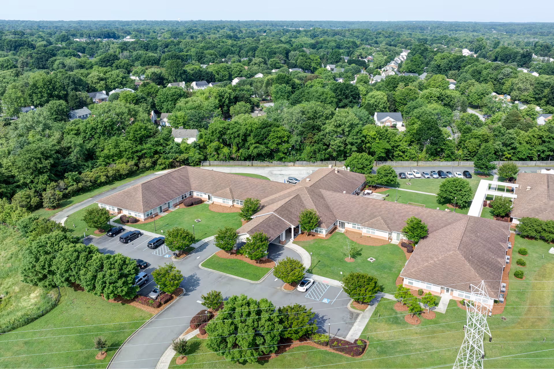 Aerial view of TerraBella Rock Hill facility showing a large, single-story building with multiple wings surrounded by green lawns, trees, and parking areas. The building has a brown roof and is situated in a suburban area with many trees and houses in the background.