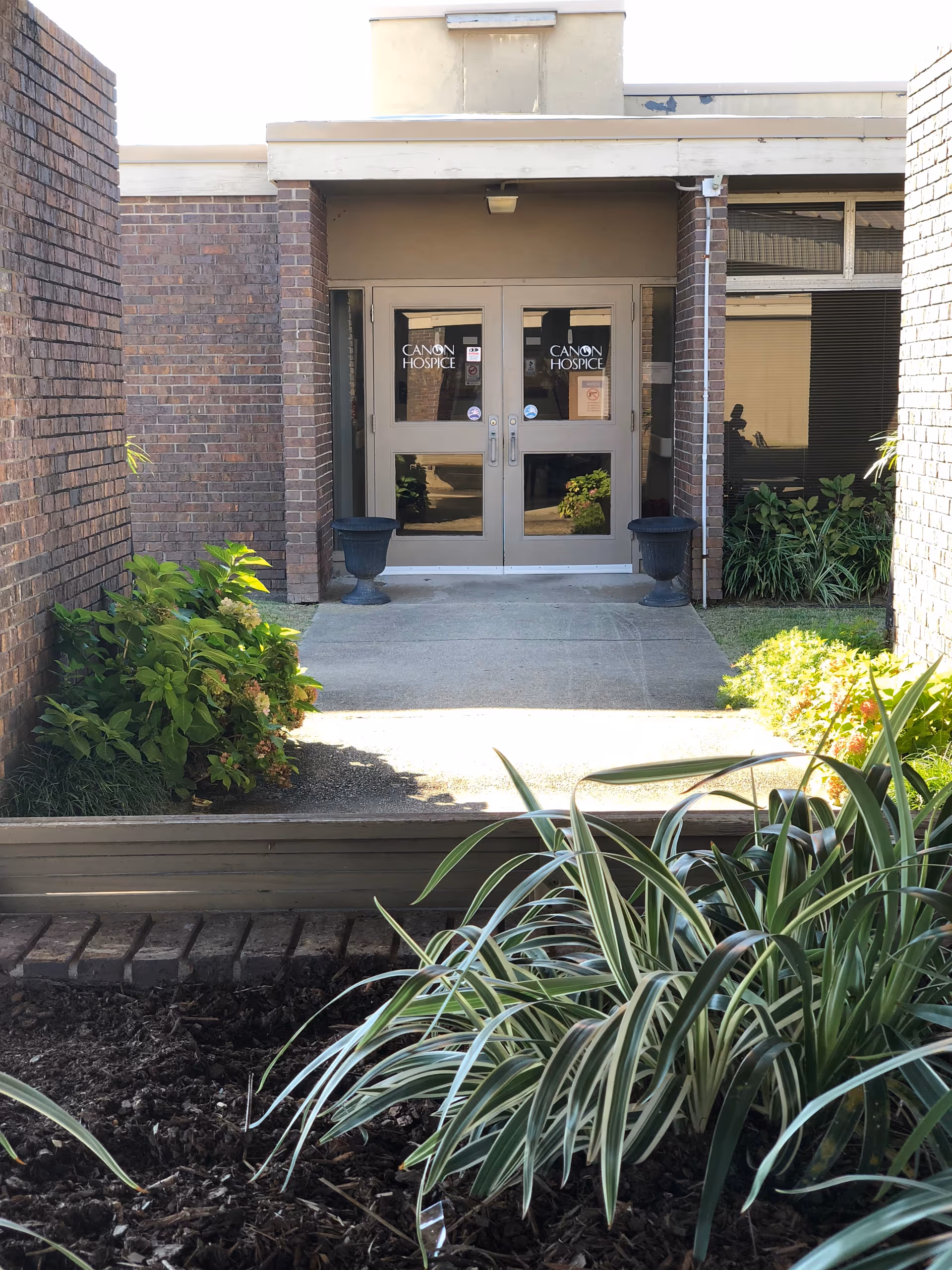 Front entrance of Canon Hospice with glass doors, brick walls, and surrounding plants.
