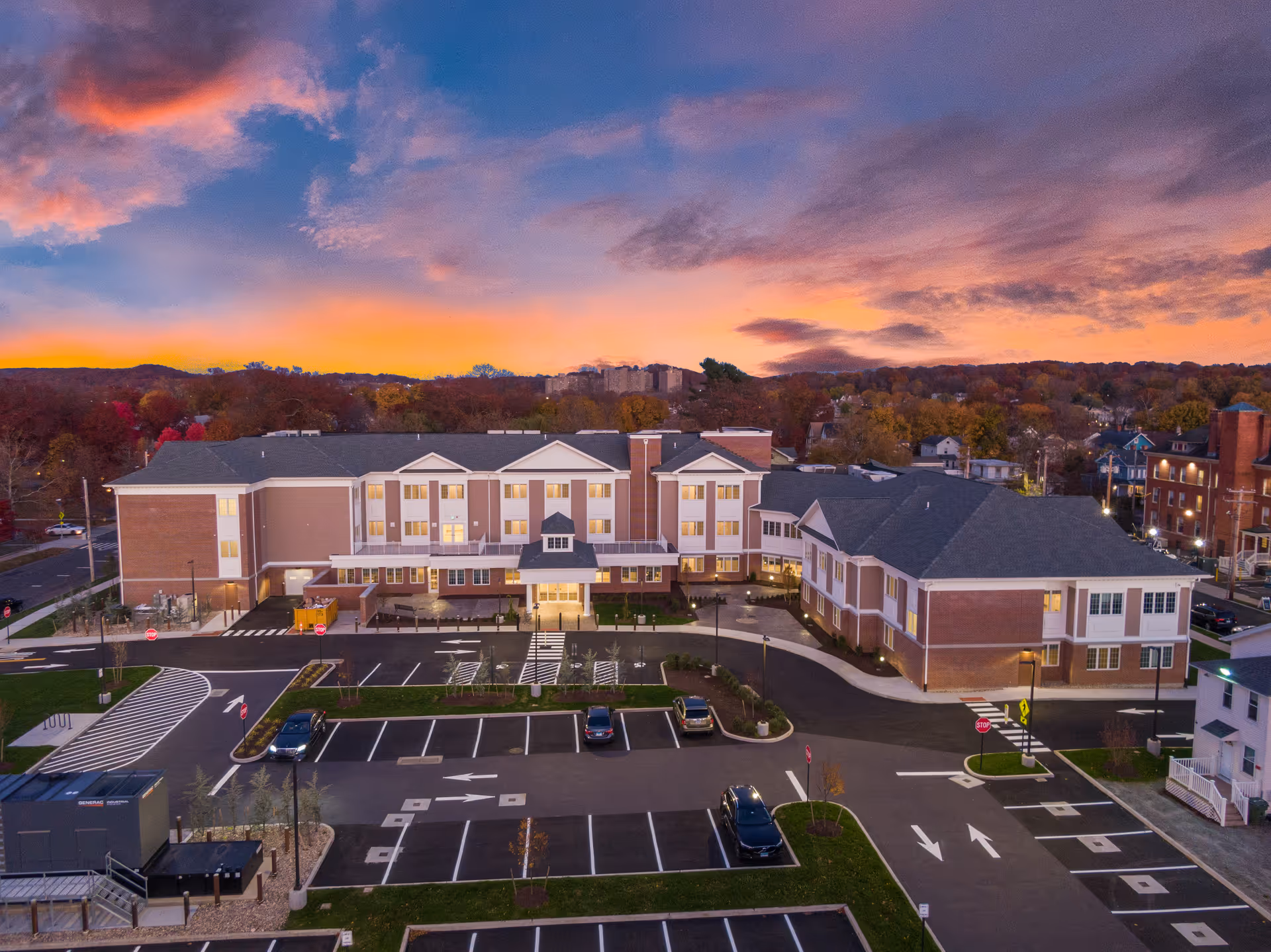 Aerial view of Chatham Place at Mary Wade senior living facility at sunset, showing a large multi-story building with a parking lot in front and a colorful sky with clouds in the background.