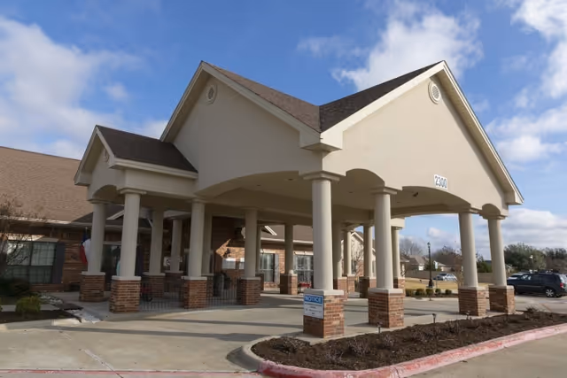 Covered entrance porte-cochere with columns at the front of a brick-and-stucco senior living facility under a blue sky.