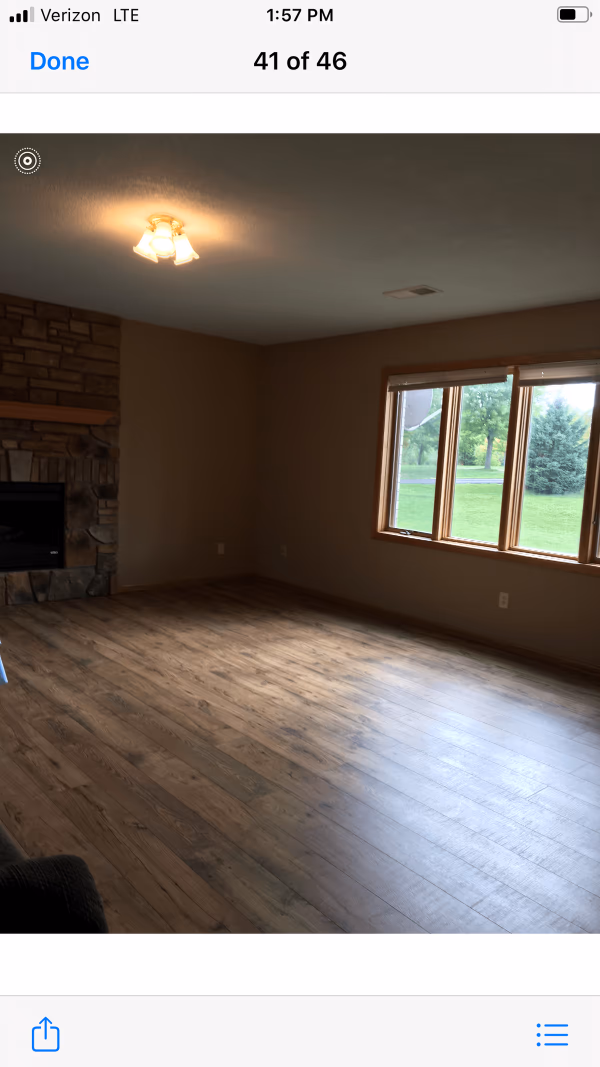 Empty living room with wooden flooring, a stone fireplace on the left wall, a ceiling light fixture, and a large window showing green grass and trees outside.