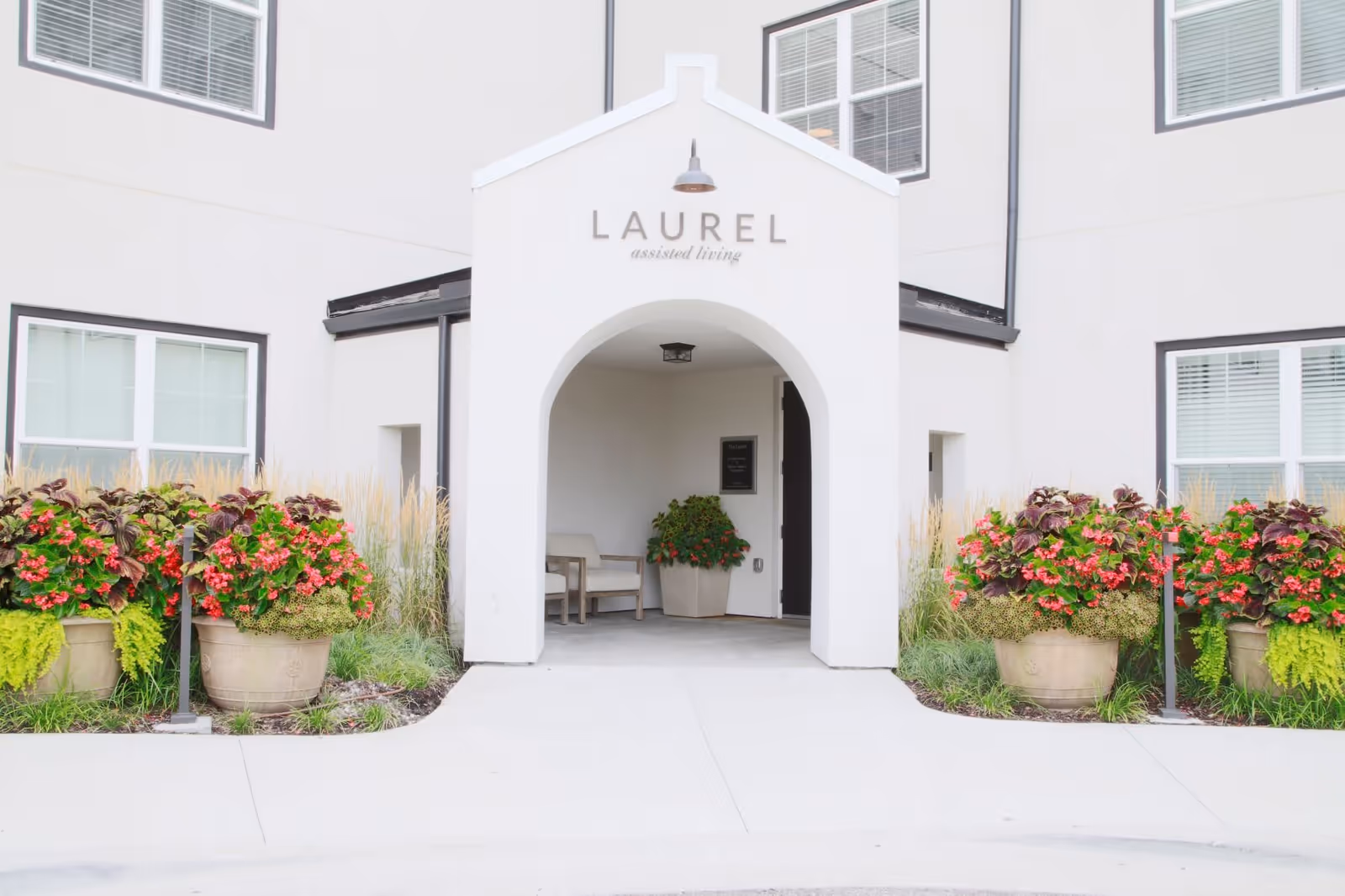 Entrance to a building with a white archway labeled 'LAUREL assisted living', flanked by large flower pots with vibrant red and green plants, and windows on the beige exterior walls.