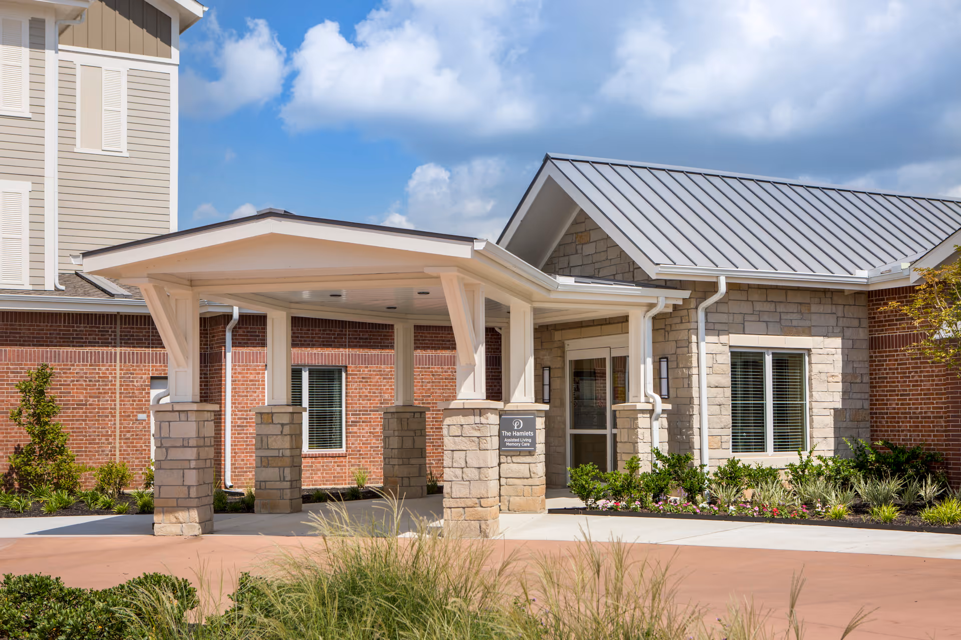 Entrance to a senior living facility with a covered drop-off area supported by stone pillars, surrounded by landscaped greenery and a clear blue sky with some clouds.