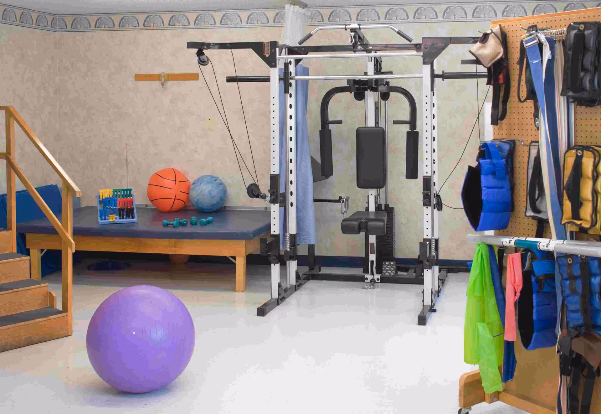 A therapy/exercise room featuring a multi-station weight machine, a raised therapy table, exercise balls, small dumbbells and hanging weighted vests.