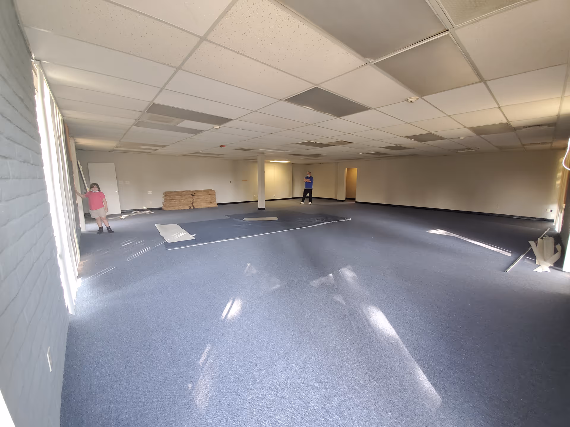 Large empty interior room with carpet, ceiling tiles, and two people standing.
