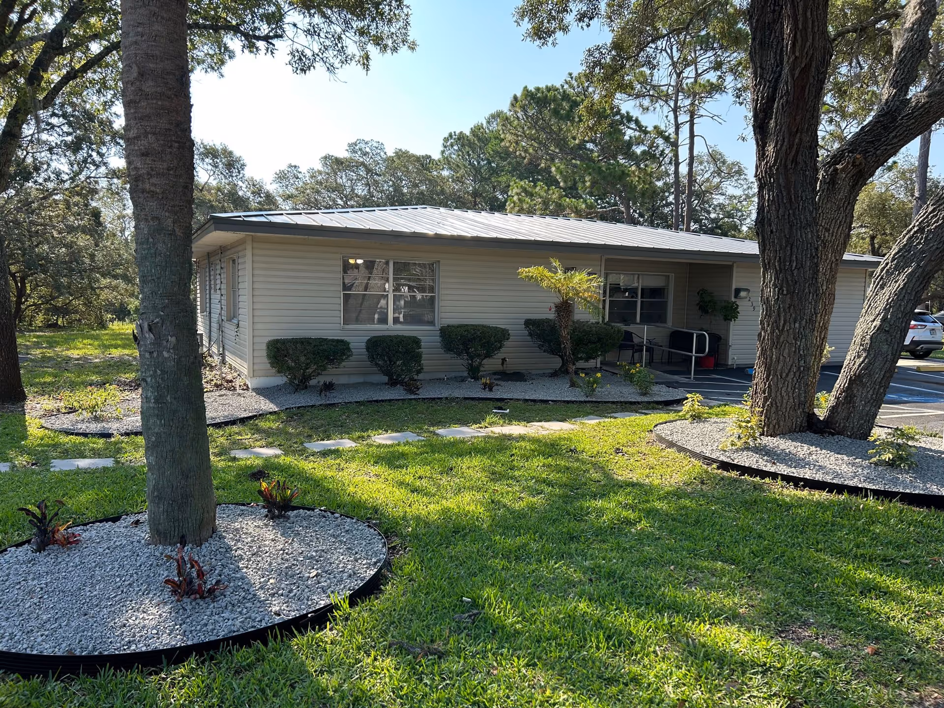 Single-story building with beige siding surrounded by green grass, trees, and landscaped areas with white gravel. There is a paved walkway leading to the entrance, which has a ramp and handrails. The sky is clear and blue.