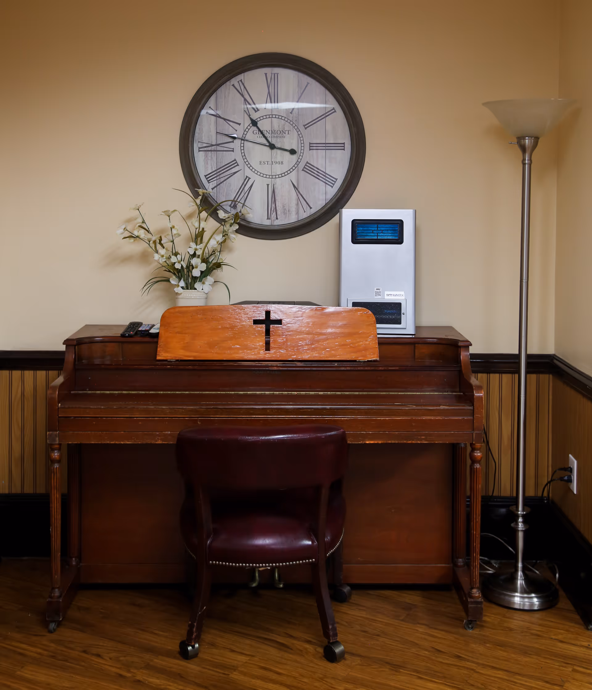 An upright wooden piano with a music stand featuring a cross cutout, a dark red chair on wheels in front of it, a vase with white flowers on top of the piano, a large round wall clock with Roman numerals above the piano, and a tall floor lamp to the right, all set against a beige wall with wood paneling at the bottom.