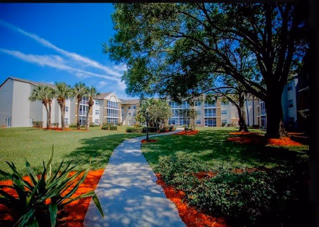 A paved walkway leads through a landscaped courtyard with grass, trees, and palm trees toward a multi-story senior living building under a blue sky.
