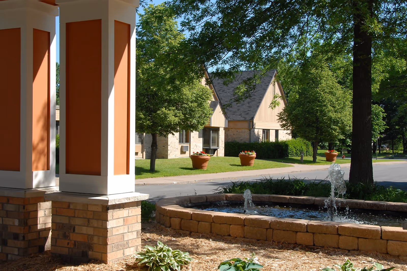 Outdoor view of a senior living facility with a small stone fountain in the foreground, green trees, large flower pots with red flowers, and beige buildings with peaked roofs in the background under a clear blue sky.