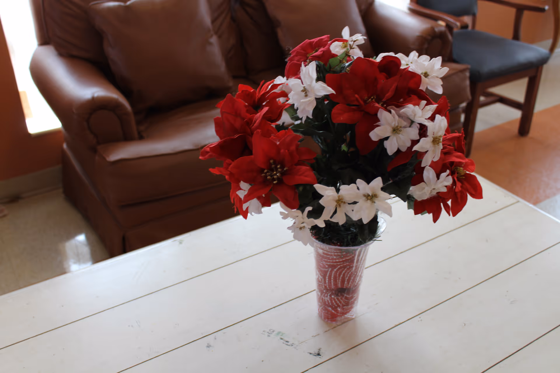 A bouquet of red and white flowers in a clear vase placed on a white wooden table. In the background, there is a brown leather armchair and a wooden chair with a dark cushion in a room with tiled flooring.