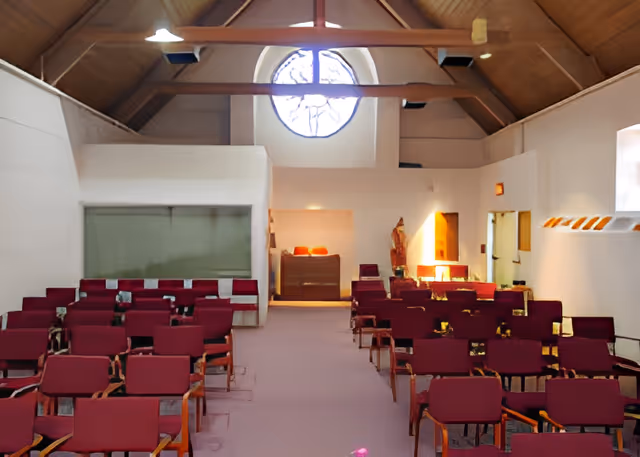 Interior view of a chapel or small auditorium with rows of red cushioned chairs arranged facing a raised area with a lectern and religious statues. The room has a high wooden ceiling with exposed beams and a circular stained glass window above the lectern area.