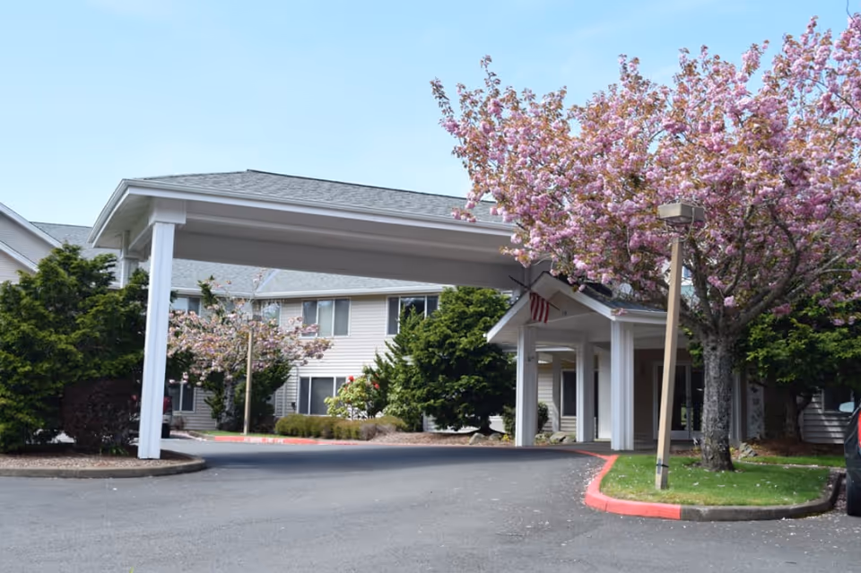 Entrance to a senior living facility with a covered drop-off area, surrounded by blooming pink cherry blossom trees and green shrubs, with a two-story beige building in the background under a clear blue sky.