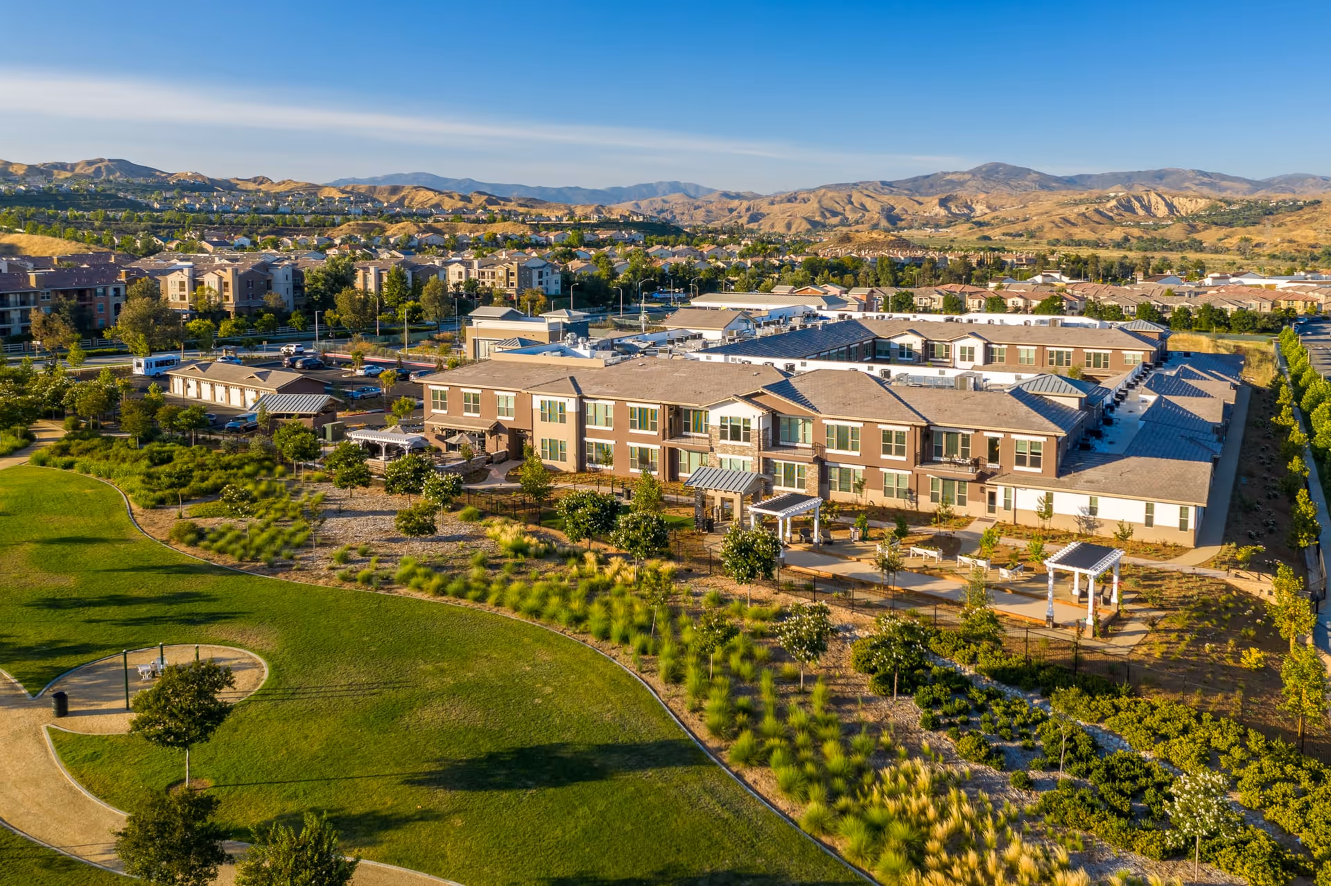 Aerial view of Oakmont of Valencia senior living facility surrounded by landscaped gardens, walking paths, and green lawns with mountains in the background under a clear blue sky.