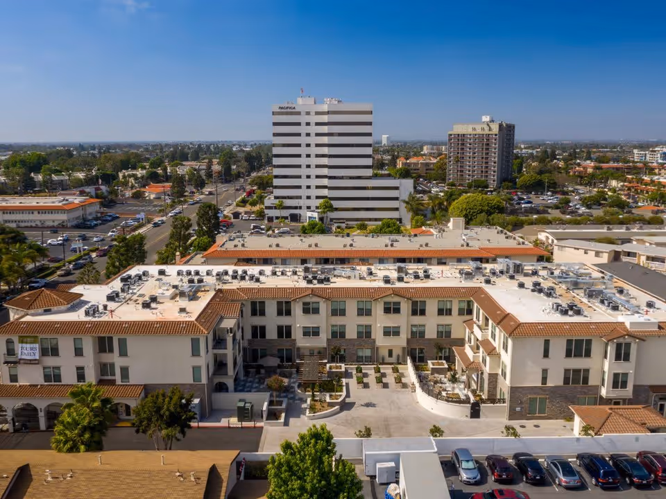 Aerial view of a multi-story senior living facility with a courtyard in the center, surrounded by a parking lot and other buildings under a clear blue sky.