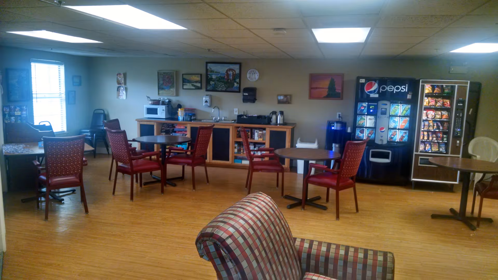 Community lounge with round tables and red chairs, a small kitchenette and vending machines in the background.
