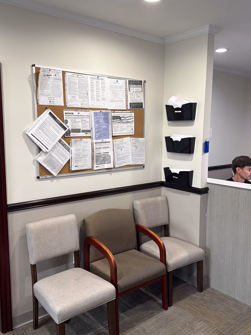 Waiting area with three chairs, bulletin board, and receptionist desk in a hallway.