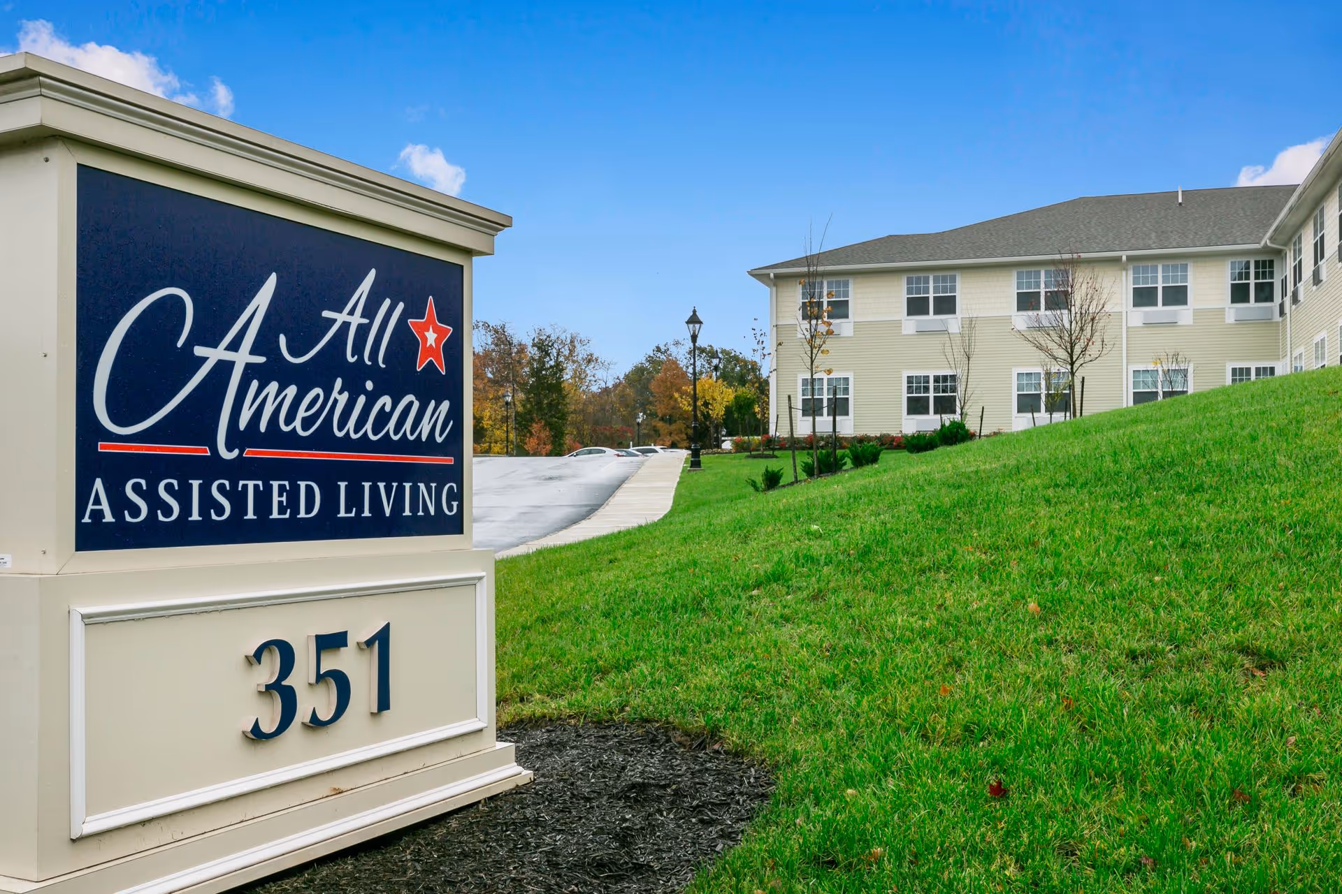Outdoor view of All American Assisted Living at Hanson facility sign with the building in the background, green grass, and a partly cloudy sky.