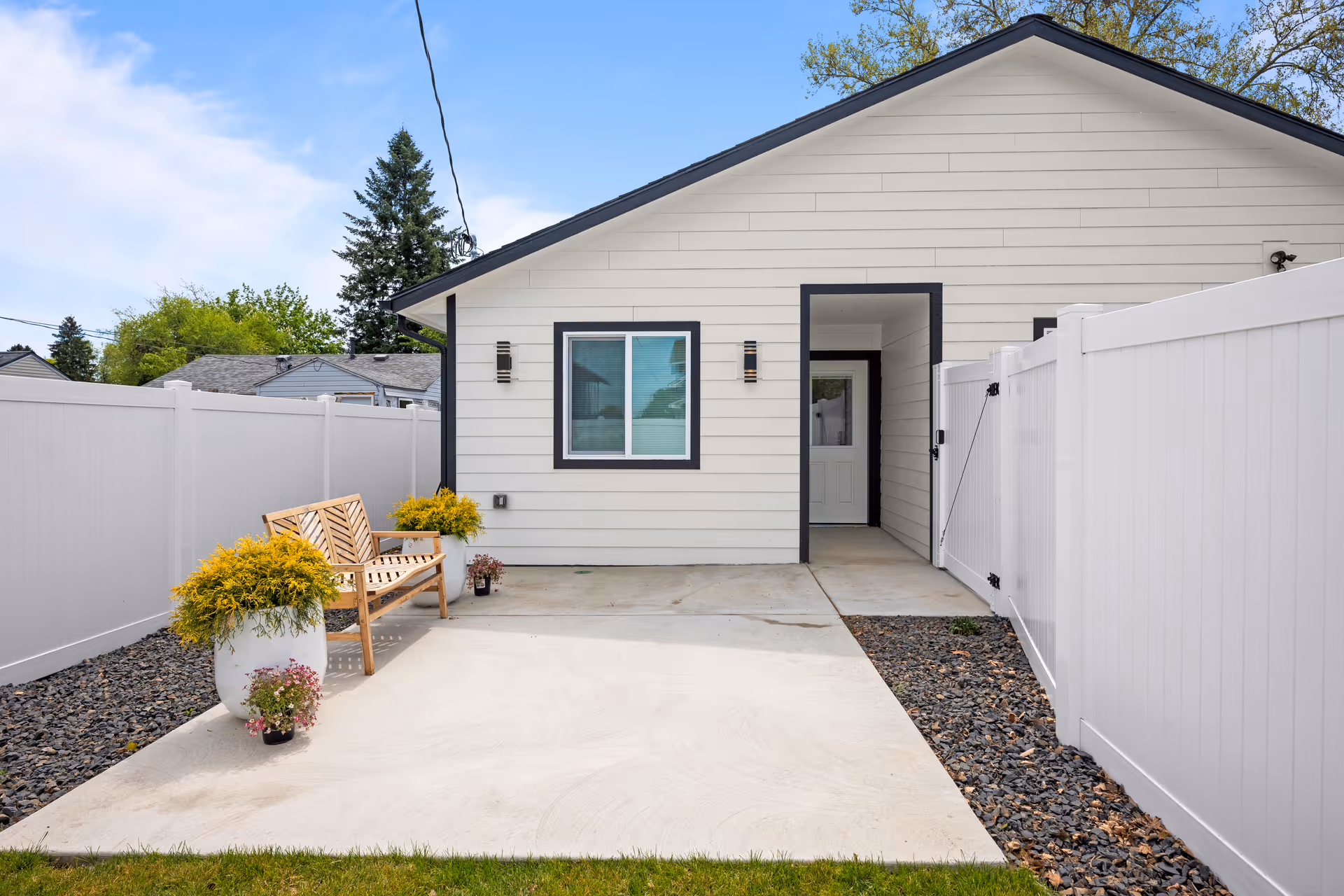 White single-story home exterior with a concrete entry path, bench, potted plants, and white fencing.